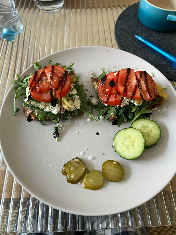 A grey plate with two toasts topped with ricotta, arugula, tomato, and balsamic glaze. Sliced cucumber and pickles are on the side, a perfect girl dinner.