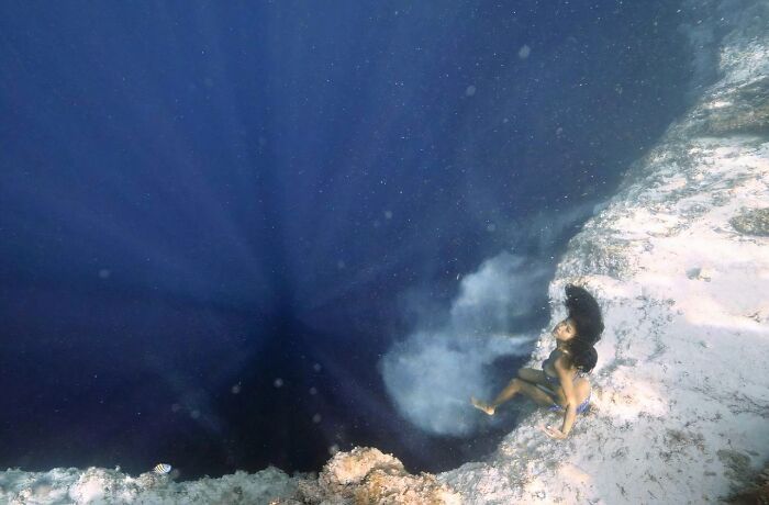 A person in a blue swimsuit underwater, near a coral reef, with deep blue water behind them. Uncomfortable water pics.