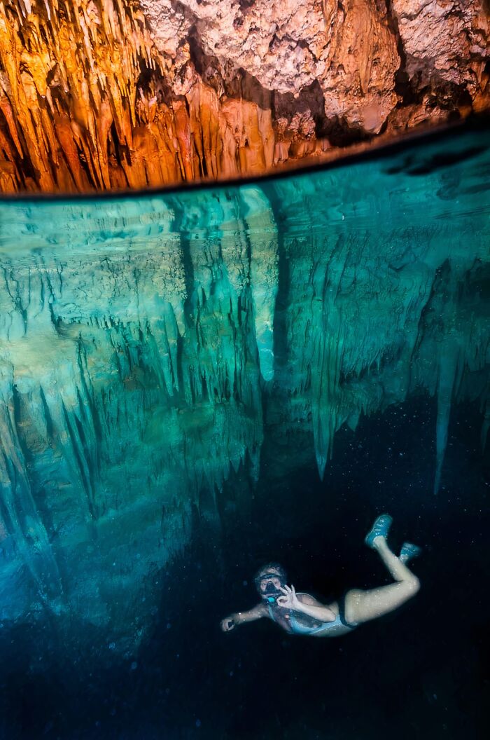 A swimmer in a mask and snorkel gives an OK sign underwater in a cave, feeling small amidst the natural water pics.
