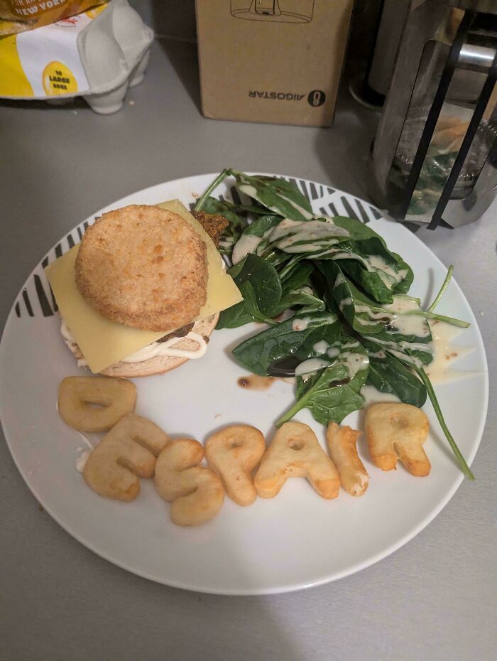 A Girl Dinner plate with a chicken burger, cheese, spinach salad, and potato letters spelling DESPAIR.