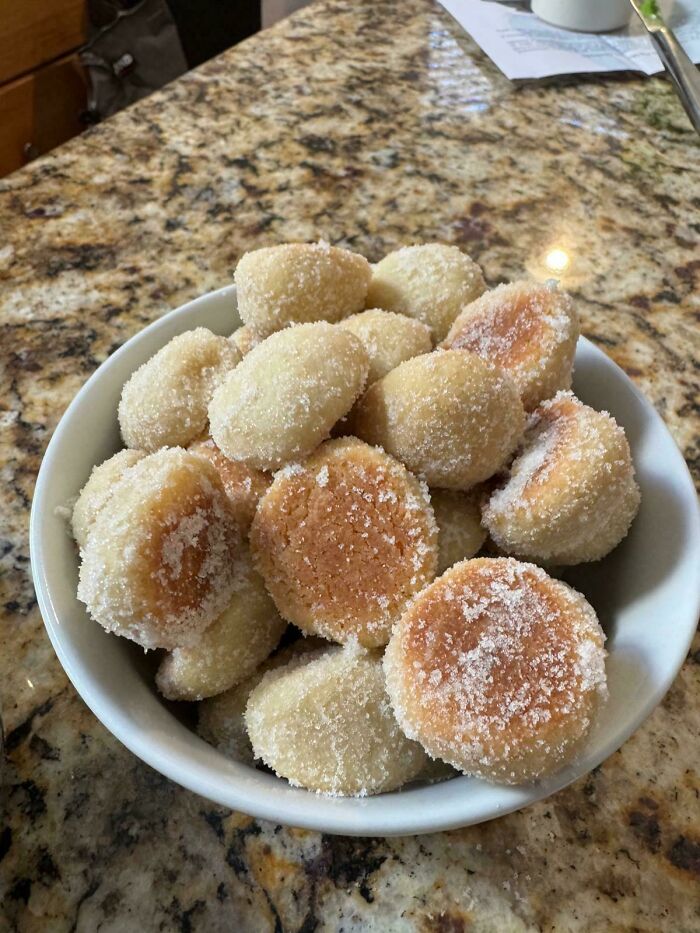A white bowl filled with Girl Dinner cookies, some lightly browned and all coated in white sugar on a granite counter.
