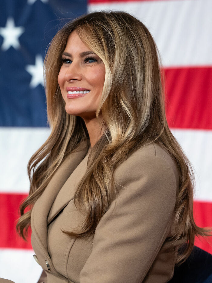 Melania Trump smiling, wearing a beige coat, with the American flag in the background. Her bio and career highlights.