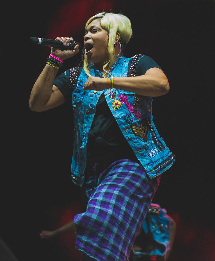 Tionne T-Boz Watkins performing live, singing into a microphone, wearing a denim vest and plaid skirt. Career highlights.