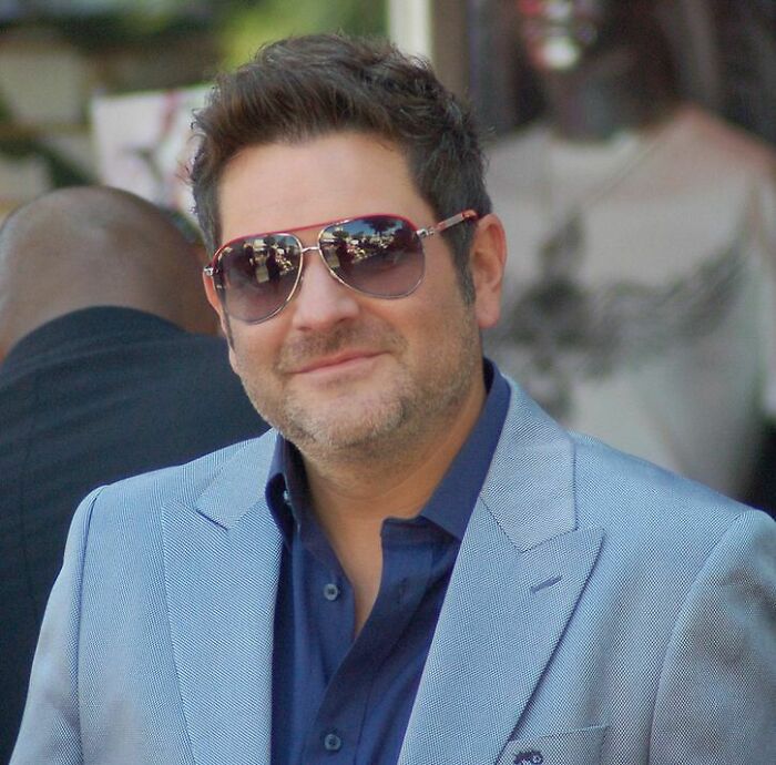 Jay DeMarcus smiling, wearing red-rimmed sunglasses, a blue shirt, and a light blue blazer. His career highlights are many.