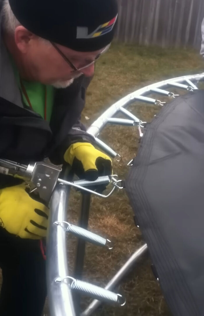 A person in yellow gloves uses a spring puller tool, showcasing a clever solution for assembling a trampoline.