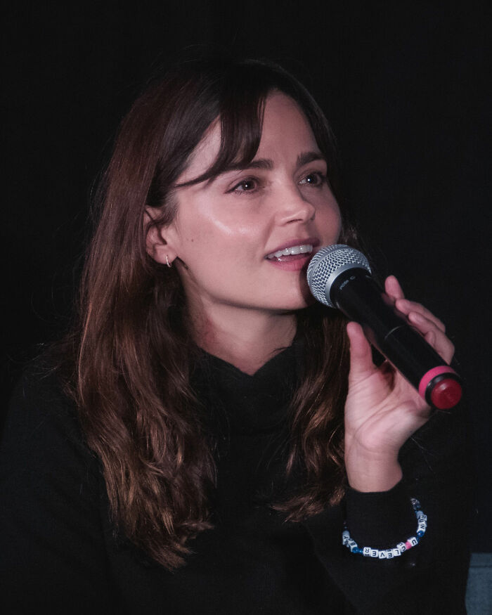 Jenna Coleman with long brown hair, speaking into a microphone at an event. This highlights her career.