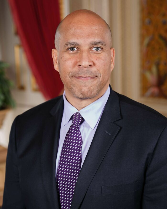 Cory Booker smiling, wearing a dark suit and purple patterned tie, posing for a portrait. His career highlights.