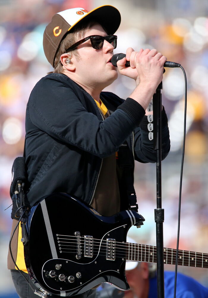 Patrick Stump performing live, wearing a cap and sunglasses, holding a microphone. His career highlights include singing and playing guitar.