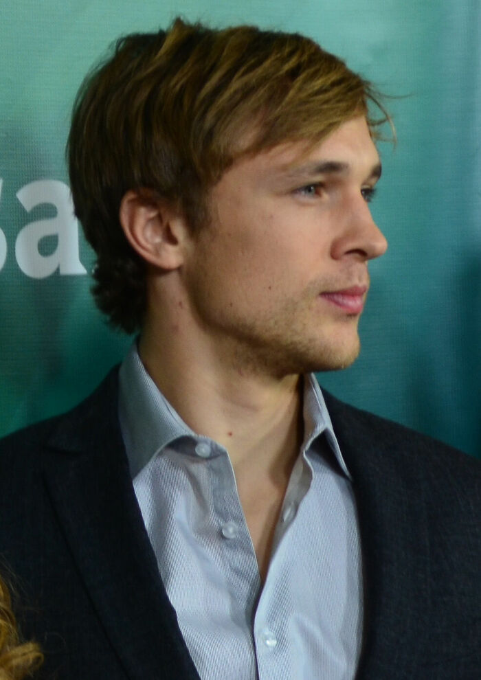 William Moseley in profile, with blonde hair and a light blue shirt under a dark jacket, at an event.