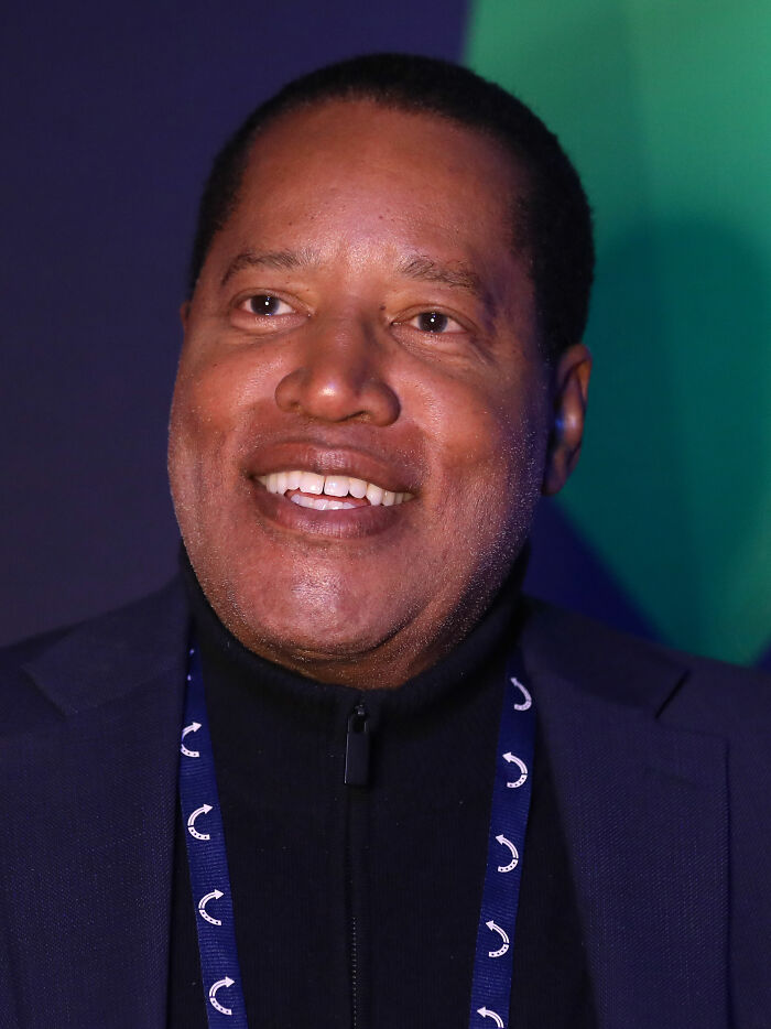 Larry Elder smiling at an event, wearing a dark suit jacket and a black turtleneck with a patterned lanyard.