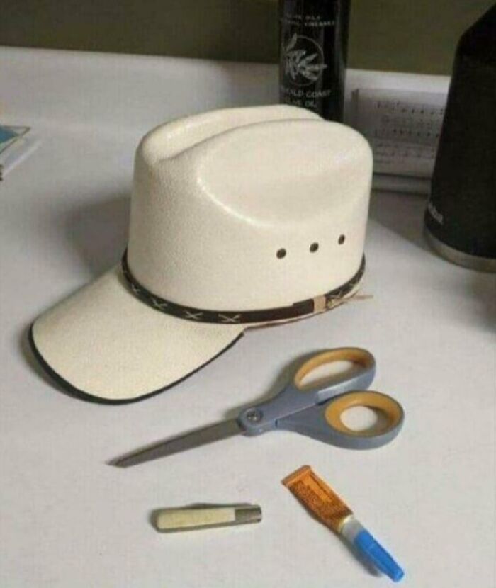 A cowboy hat converted to a baseball cap with scissors, knife, and glue on a table, a DiWHY craft gone wrong.