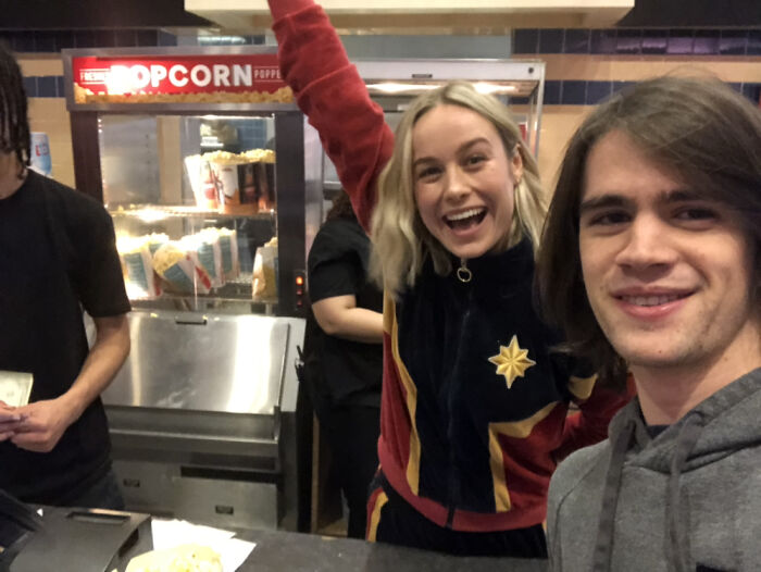 A fan taking a selfie with Brie Larson, who smiles widely at a movie theater popcorn counter. Wild photo of a celebrity encounter.