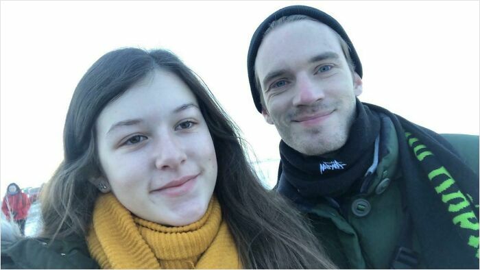 A young woman in a yellow scarf and a man in a black beanie, resembling PewDiePie, smiling for a selfie, a wild celebrity encounter.