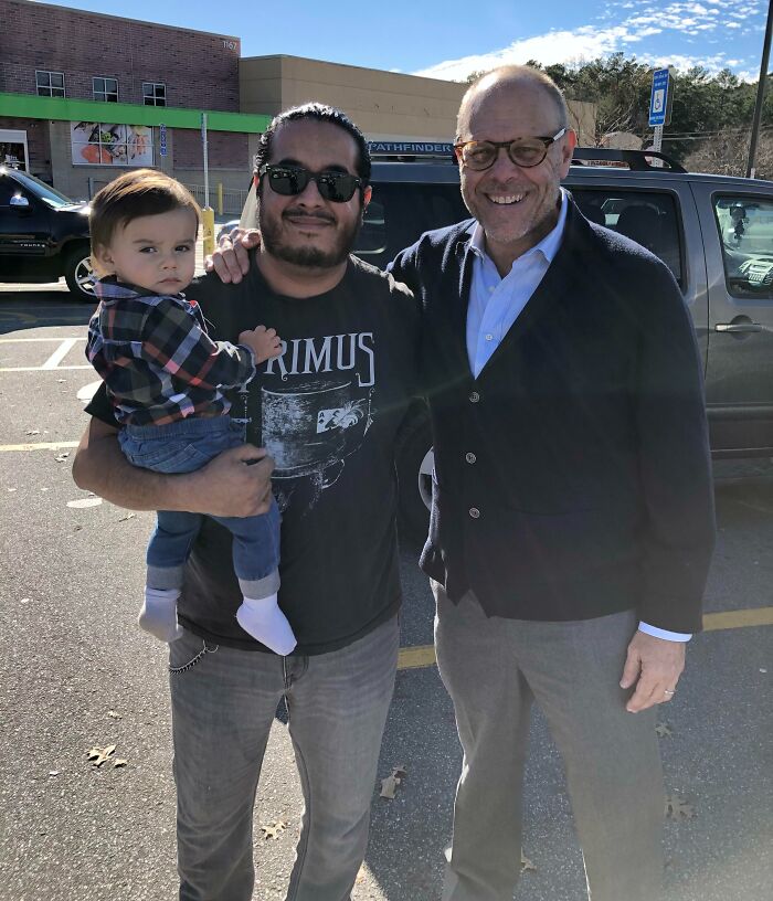 A man holds a baby while posing with a celebrity wearing glasses and a cardigan, all smiling for the camera. Wild celebrity encounters.