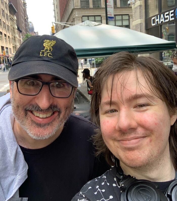 A smiling John Oliver in a black LFC cap with a fan, capturing a fun celebrity encounter moment.