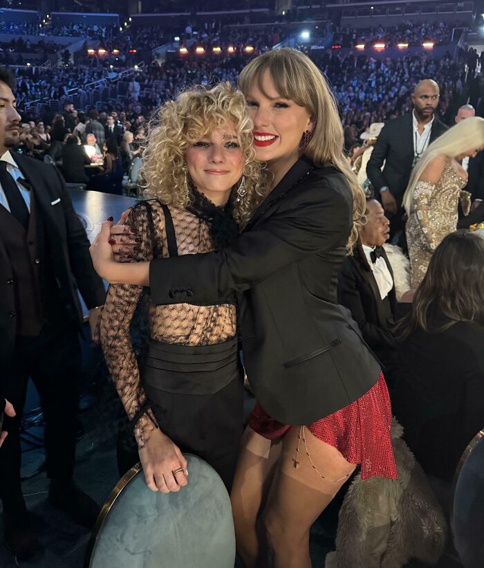 Taylor Swift in a black blazer and red skirt hugging a fan with curly hair. A memorable celebrity encounter.
