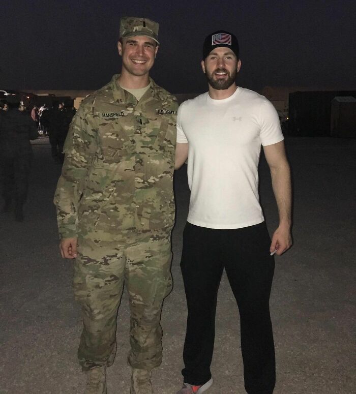 A soldier in uniform stands next to actor Chris Evans in a celebrity encounter photo. Evans wears a white t-shirt, black pants, and a US flag hat.