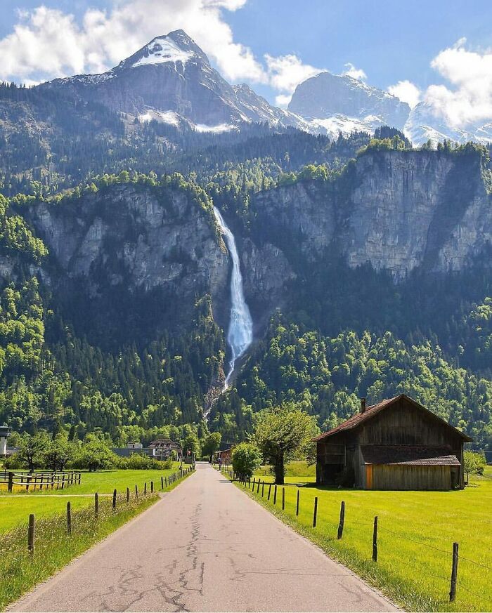 A stunning waterfall cascades down a mountain, with a serene valley, road, and rustic house below, a gentle reset for your day.