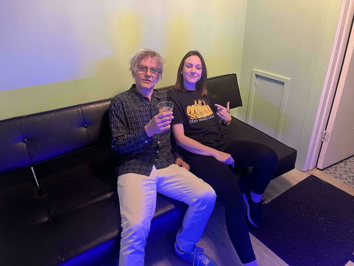 A man and woman sit on a black sofa. He holds a drink; she points at her Dean Wareham shirt. Celebrity encounters.