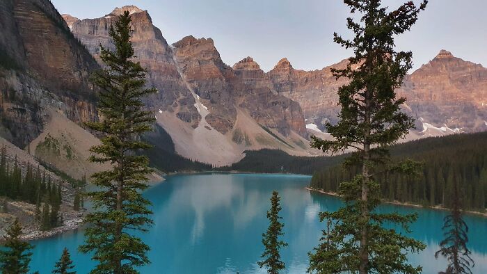 Majestic mountains frame a tranquil turquoise lake with pine trees in the foreground, offering a satisfying visual reset.