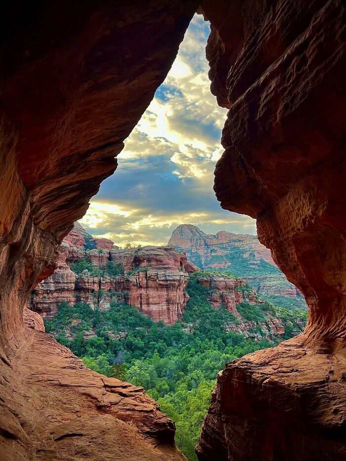 A dramatic view from inside a cave, revealing red rock canyons and green trees under a cloudy sky. Satisfying picture.