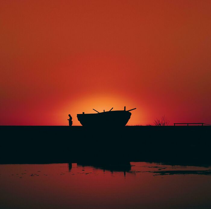 A satisfying picture of a vibrant orange sunset silhouette of a lone figure next to a boat by still water. Gentle reset.