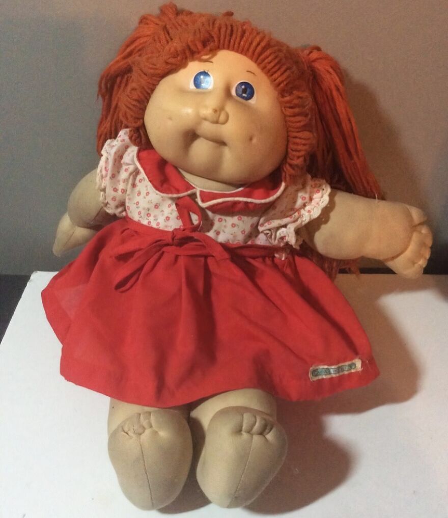 A Cabbage Patch Kid doll with red yarn hair, wearing a white floral shirt and red dress, a trip down memory lane for many.