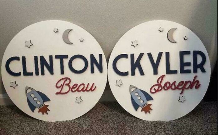 Two round, white wooden signs with space-themed cutouts. The left sign reads "CLINTON Beau," the right "CKYLER Joseph," reflecting parents naming decisions.