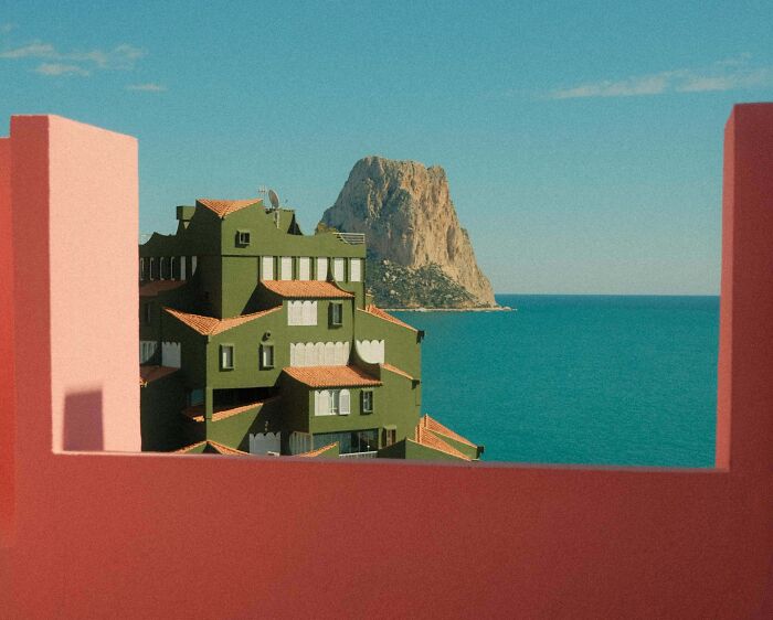 A unique green building with terracotta roofs by the sea, framed by pink and red walls. Impressive architecture.