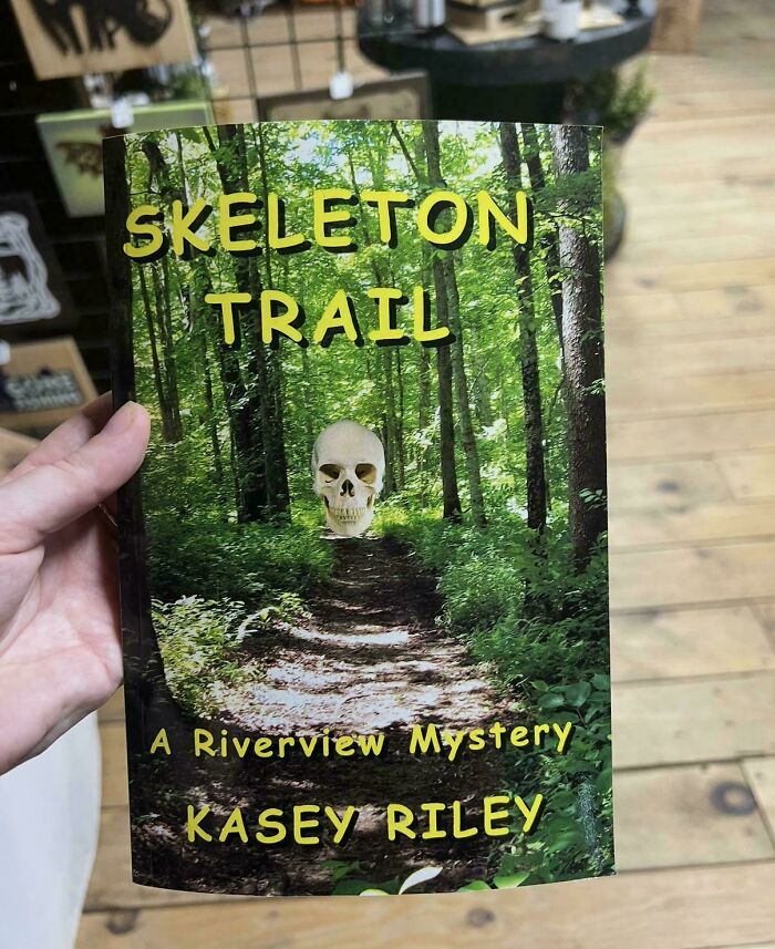 A hand holding a book cover featuring a skull over a forest path, showcasing unhinged book covers.
