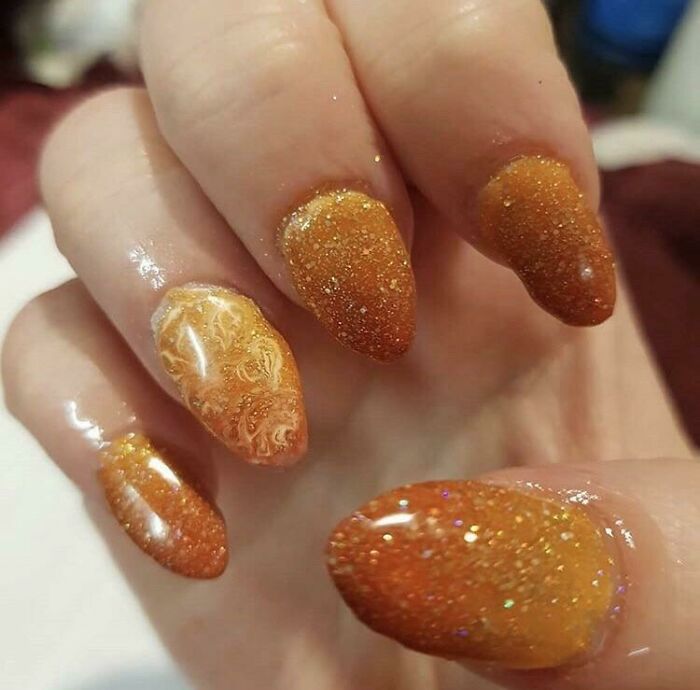 Close-up of orange, glittery nails with one patterned nail. A potential beauty disaster from the salon.