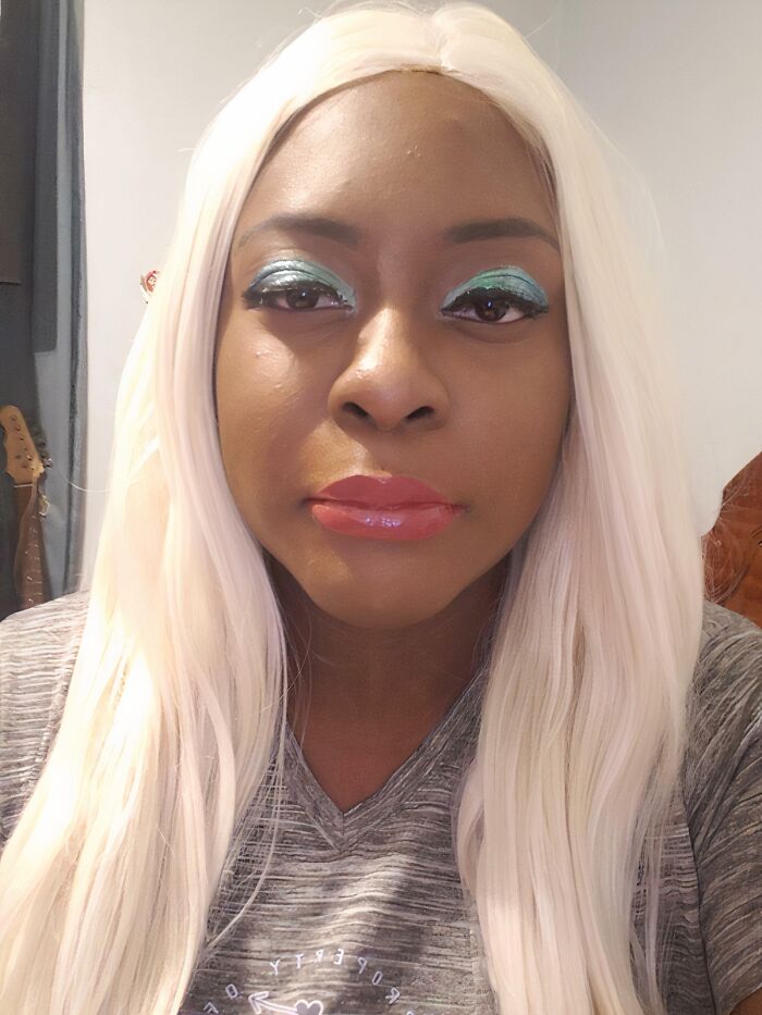 A woman with platinum blonde hair and teal eyeshadow has a beauty disaster: an uneven, glossy red lip.