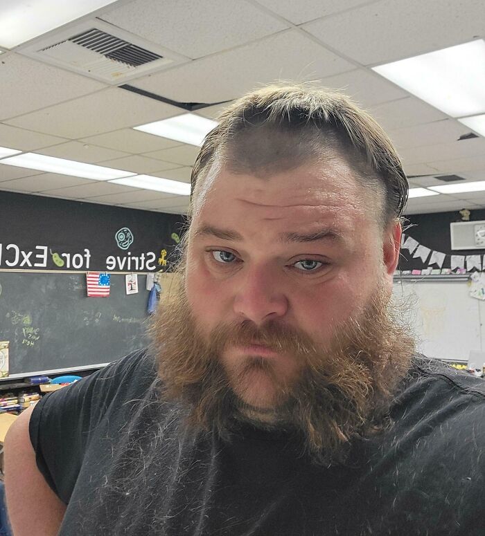 A man with a serious expression and a bad haircut, a true beauty disaster, in a classroom setting.