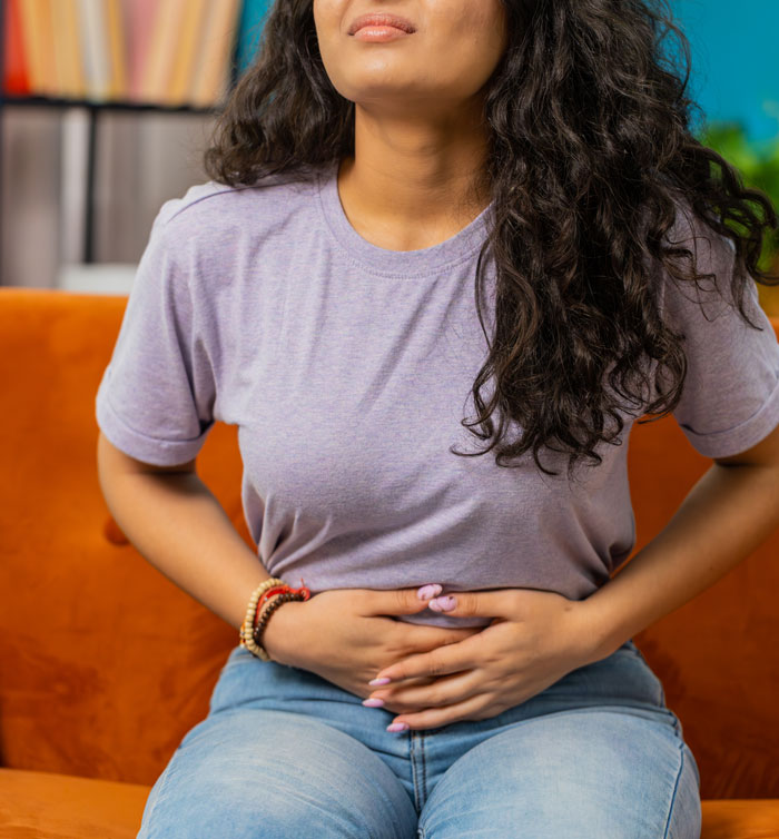 Woman sitting on orange couch holding stomach, expressing discomfort, related to things women are tired of explaining to men.