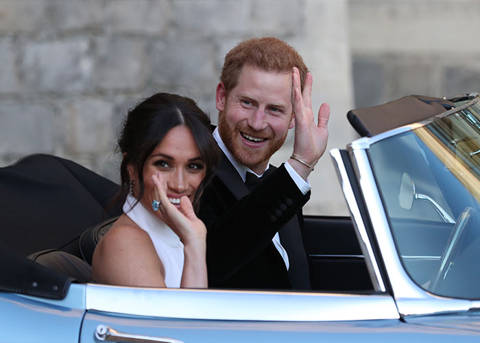 Prince Harry and Meghan Markle smiling and waving from a vintage convertible car during a public appearance