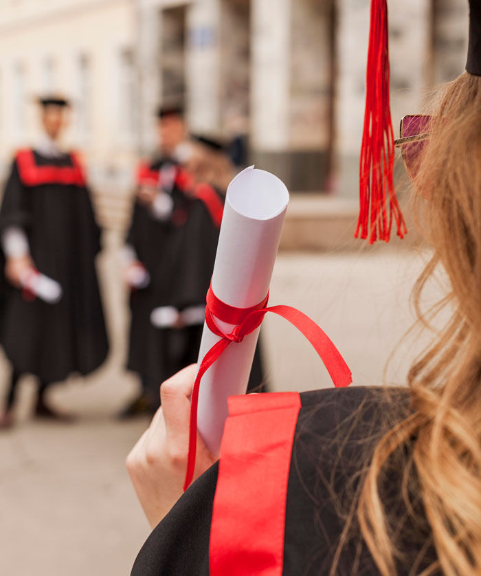 Graduate holding diploma with red ribbon, wearing cap and gown, celebrating a milestone achievement outdoors.