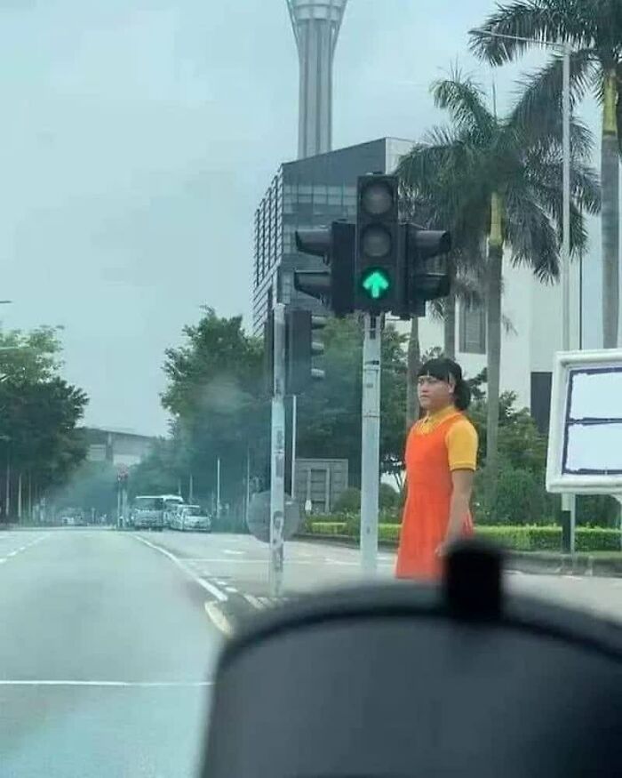 A weird image of a person dressed as the Squid Game doll stands next to a traffic light.