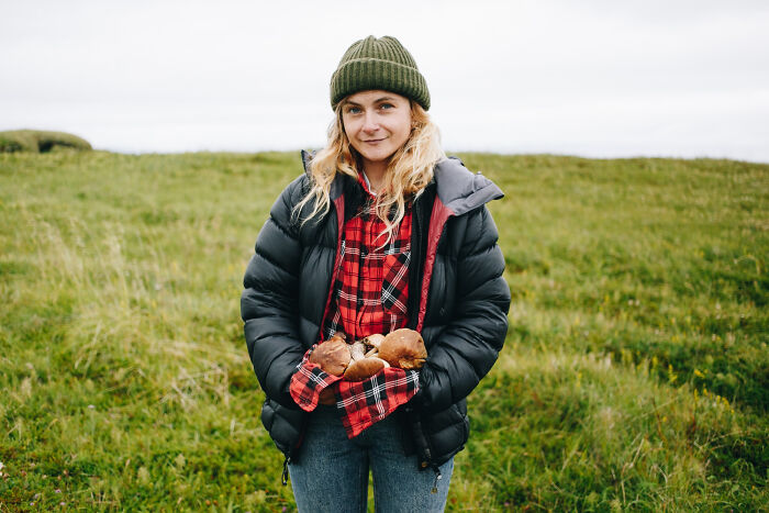 A woman in a green beanie and black puffer jacket holding mushrooms, against a green field. Wildest rumors and a lot to process.