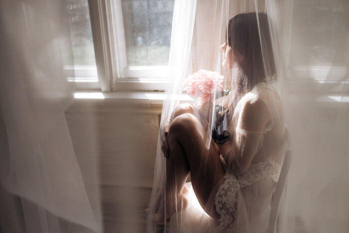 Bride sitting by window in a sheer veil, holding a bouquet, reflecting on future breakup signs at a wedding event.