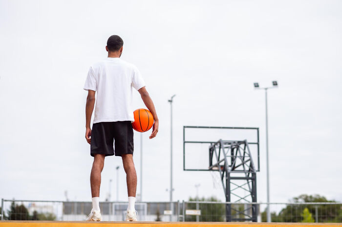 Rear view of a basketball player holding a ball on a court. It depicts the personal costs of being a celebrity relative.