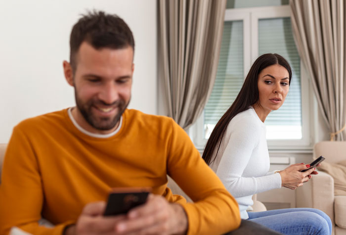 Uma mulher olha com desconfiança para o marido, que sorri enquanto usa o telefone, suspeitando que ele tenha um filho secreto.