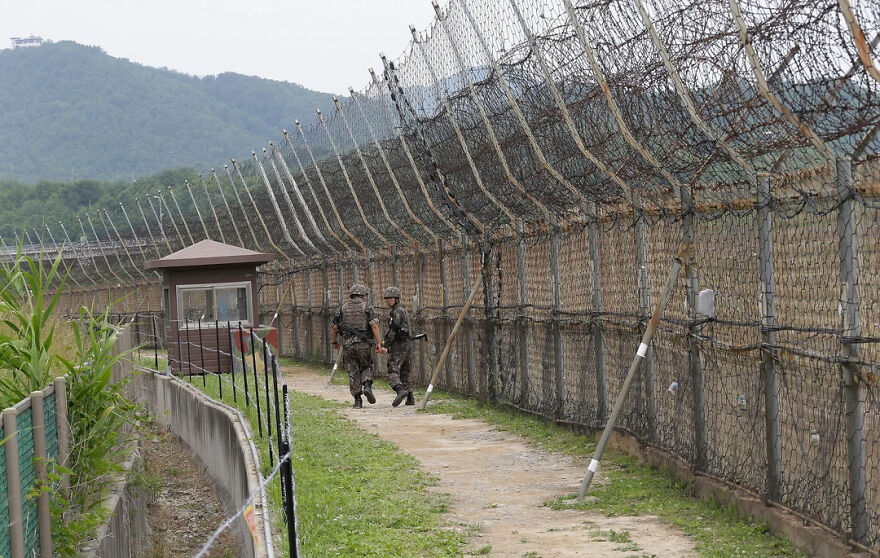 Two soldiers patrol along a formidable security fence with coiled barbed wire, a guard shack visible, highlighting tough security.