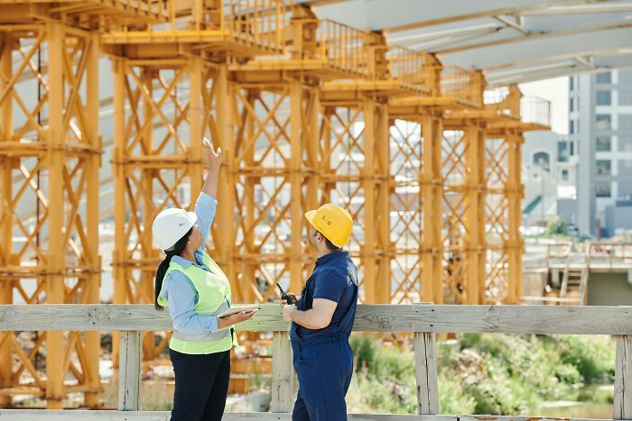 Two construction workers, one pointing, wearing hard hats at a construction site. It highlights potential workplace incidents.