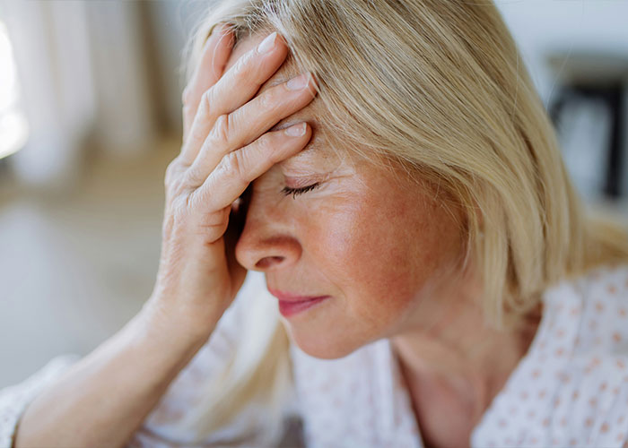 Uma mulher loira angustiada segurando a testa e os olhos fechados, possivelmente experimentando um efeito colateral relacionado ao tratamento de Alzheimer.