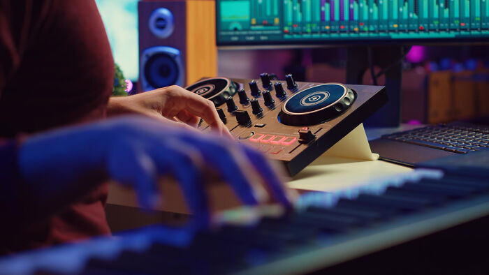 A person's hands on a DJ mixer and keyboard, with a computer screen displaying audio software in the background. Celebrity relatives.