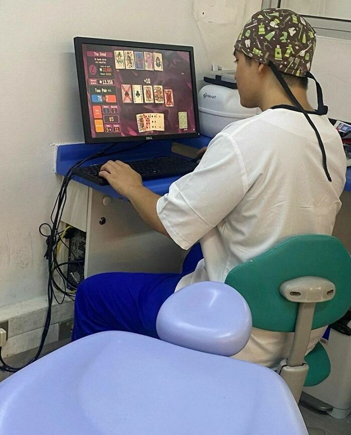A healthcare worker in scrubs and a dental-themed scrub cap plays a card game on a computer. A weird image, indeed.