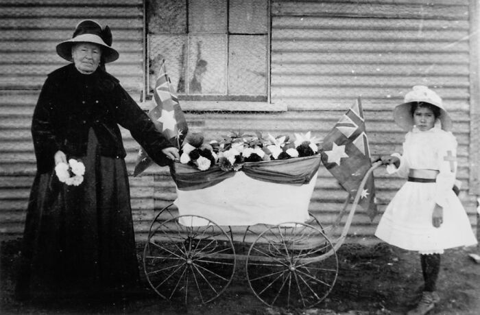 Black and white rare photo of an elderly woman and girl with decorated vintage pram holding flags outdoors.