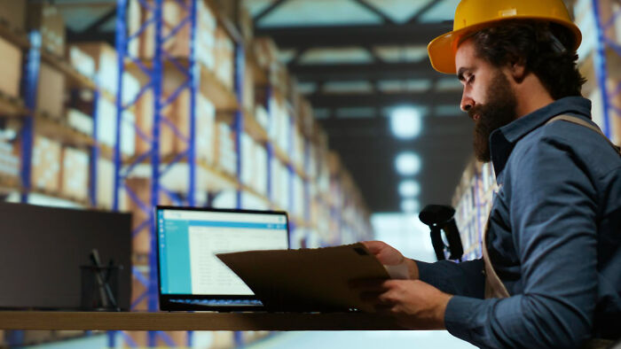 Man wearing hard hat reviews documents at desk with laptop in warehouse, illustrating jobs AI can’t replace in construction.