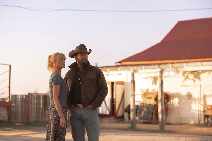 A man in a cowboy hat and a woman stand outside a building, related to Dutton Ranch show drama.
