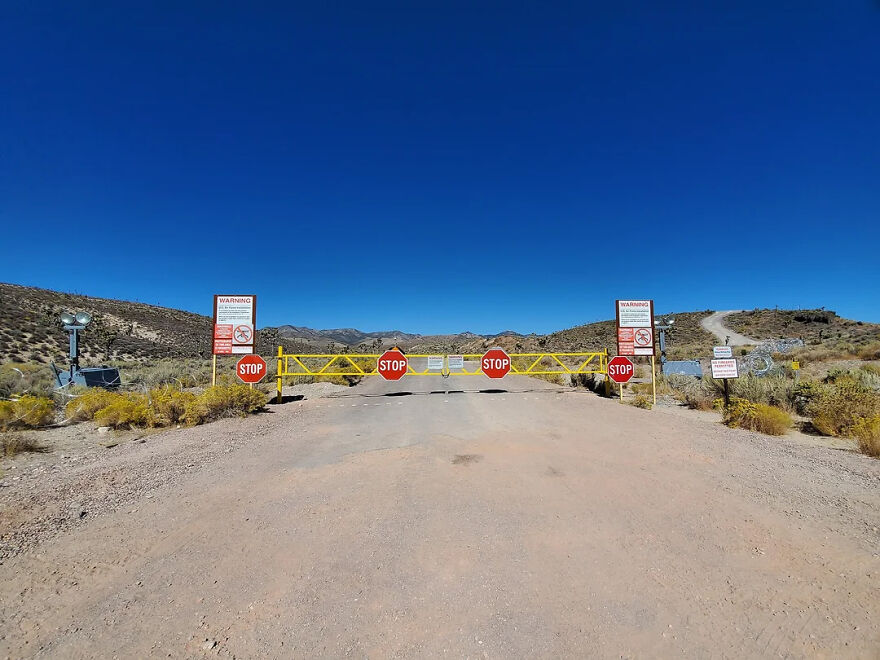 A dirt road blocked by a yellow gate with four red stop signs, warning of toughest security ahead.